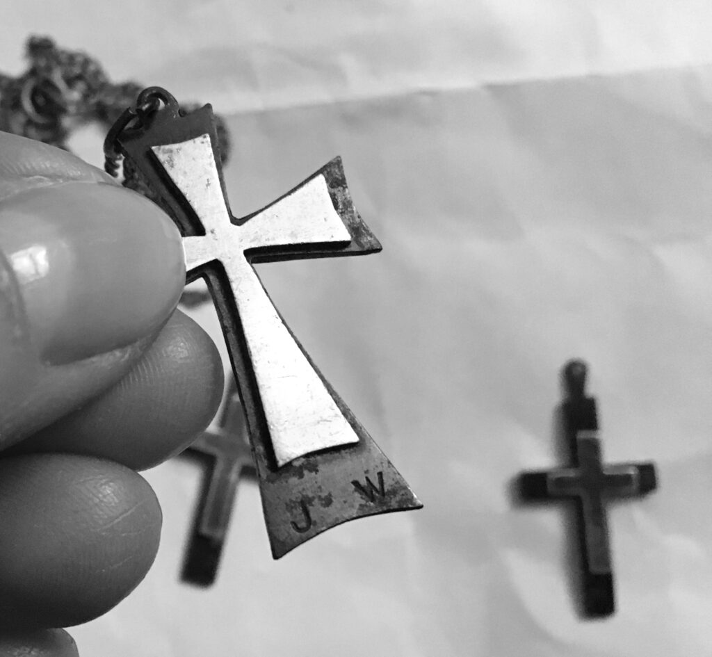 Zdjęcie czarno-białe. W kobiecych dłoniach mały metalowy krzyżyk. U podstawy widać inicjały J.W. W dalszym planie dwa mniejsze krzyżyki. 