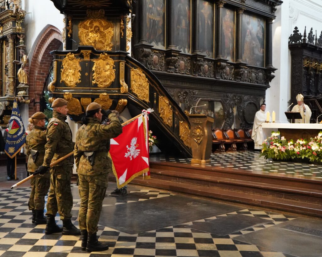Na zdjęciu poczet sztandarowy wojska polskiego. Żołnierze 2 panie, jeden pan ubrani w mundury moro. Kobiety salutują przed duchownym stojącym za ołtarzem, mężczyzna pochyla sztandar. 