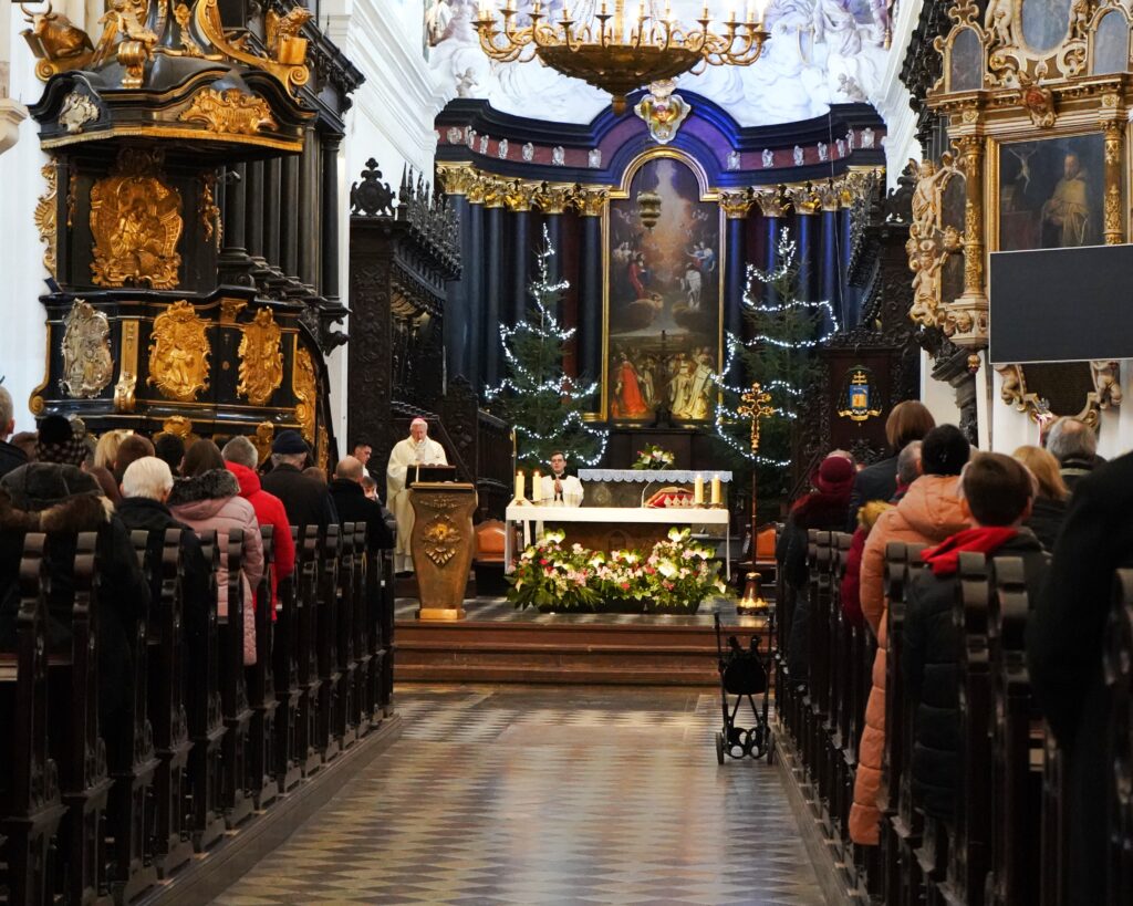 Na zdjęciu wnętrze kościoła podczas uroczystości. W ławkach widać ludzi ubranych w grube kurtki. W oddali na tle ołtarza duchowny ubrany w białe szaty liturgiczne odprawia nabożeństwo. 
