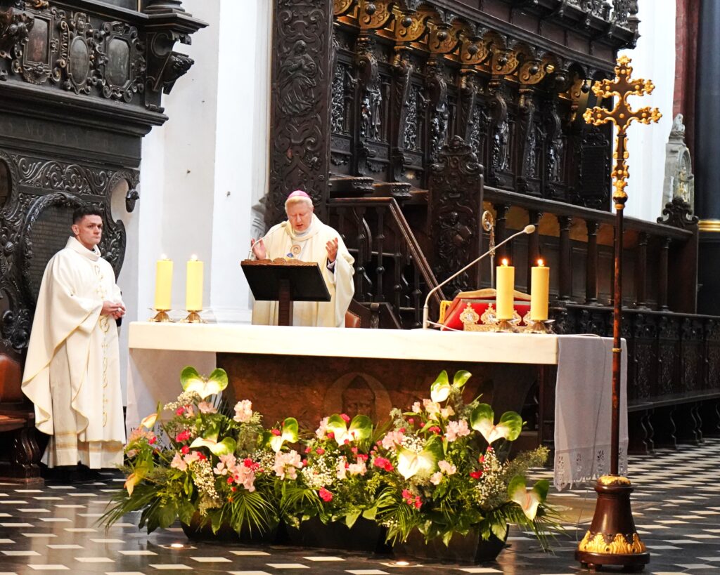 Na zdjęciu przed ołtarzem dwóch duchownych. Ubrani w białe szaty liturgiczne. Stojący po lewej stronie starszy duchowny w szatach biskupich wznosi swoje dłonie do góry. Z tyłu widoczne bogato zdobione ławy kościelne. 