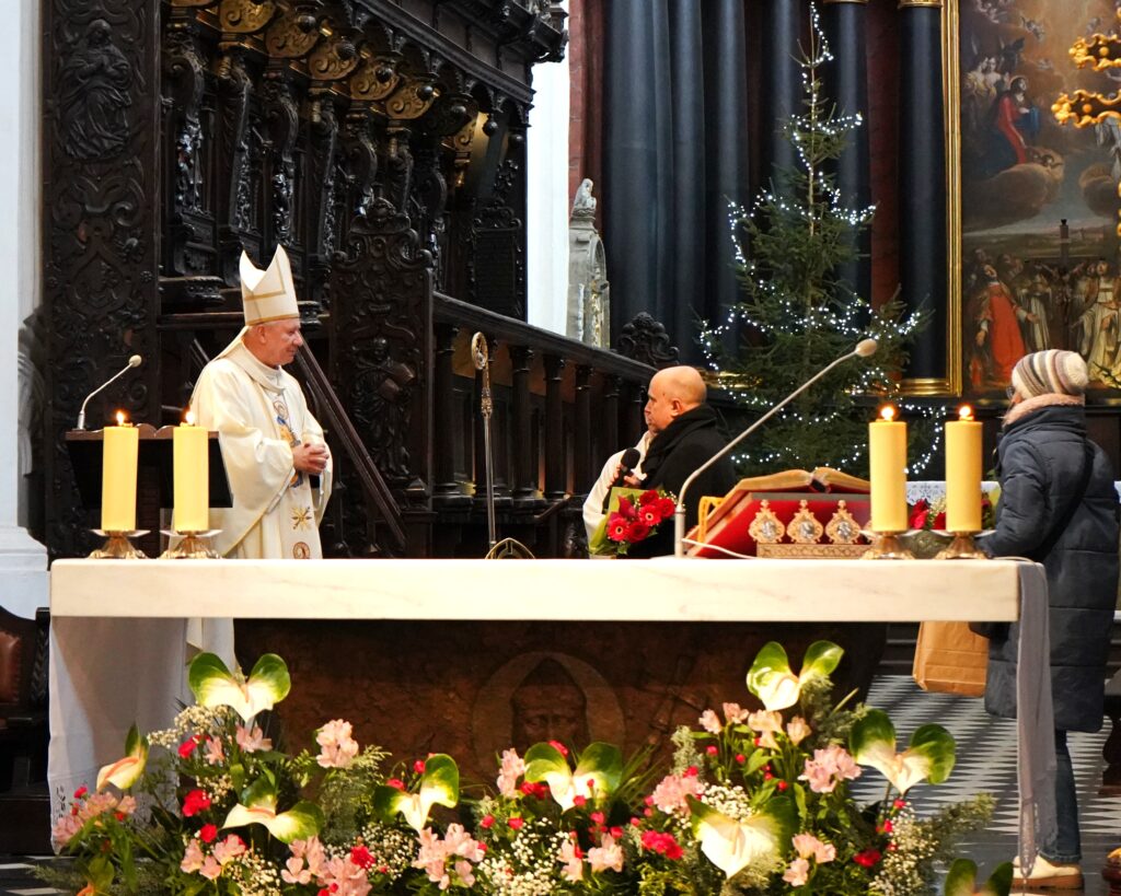 Na zdjęciu za ołtarzem stoją trzy postacie. 2 mężczyzn i jedna kobieta. Z prawej strony ubrany w białe szaty liturgiczne i biskupia mitrę duchowny. Po środku mężczyzna ubrany w czarny płaszcz, czarny szalik mężczyzna trzyma w dłoniach kwiaty i przemawia do mikrofonu. Z lewej strony ubrana w ciemno niebieską kurtkę i grubą czapkę kobieta. 