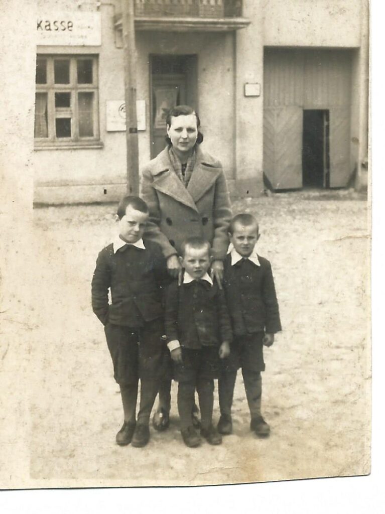 Na czarno-białym zdjęciu kobieta z trójką dzieci - chłopców. Ubrani elegancko, kobieta średniej długości włosy, szary płaszcz. Chłopcy krótko ostrzyżeni, czarne stroje z których wystają białe kołnierze. Z tyłu widać budynek z częściowo widocznym napisem po niemiecku kasie. 