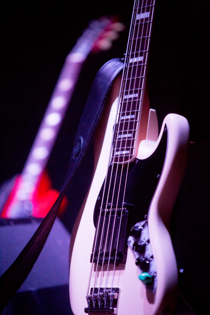 Na zdjęciu podświetlona biała gitara basowa stojąca na statywie. 