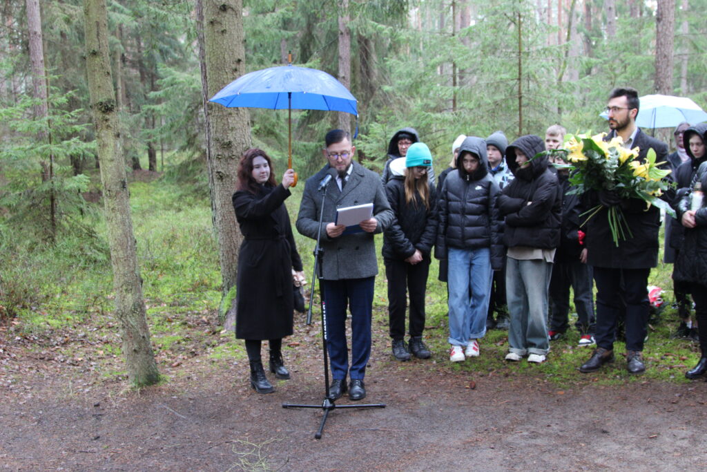 Na zdjęciu widzimy grupę osób zgromadzoną w lesie. Jedna osoba przemawia, a inna trzyma nad mówcą parasol. Wygląda to na poważne lub ważne wydarzenie na świeżym powietrzu. Zdjęcie jest interesujące ze względu na nietypowe miejsce takiego zgromadzenia i jedność ludzi w naturalnych warunkach.