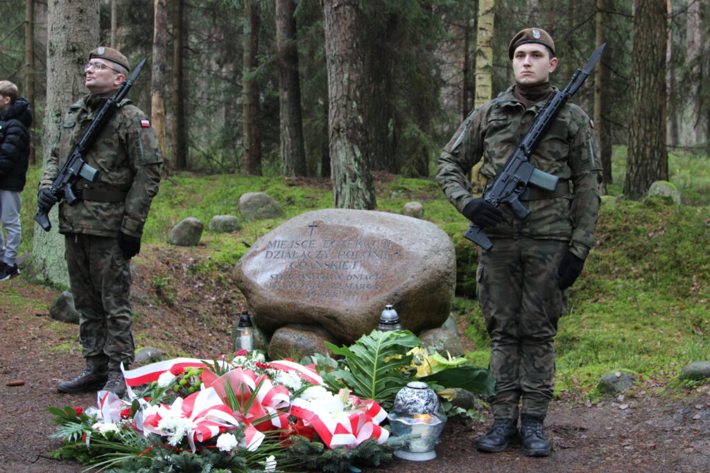 Na zdjęciu widoczny kamień upamiętniający ofiary egzekucji z 22 marca 1940 r. Przed kamieniem warta honorowa. Po obu stronach żołnierze ubrani w  mundury moro. Przed pomnikiem ułożone wieńce z biało-czerwonymi szarfami. W oddali widać las. 