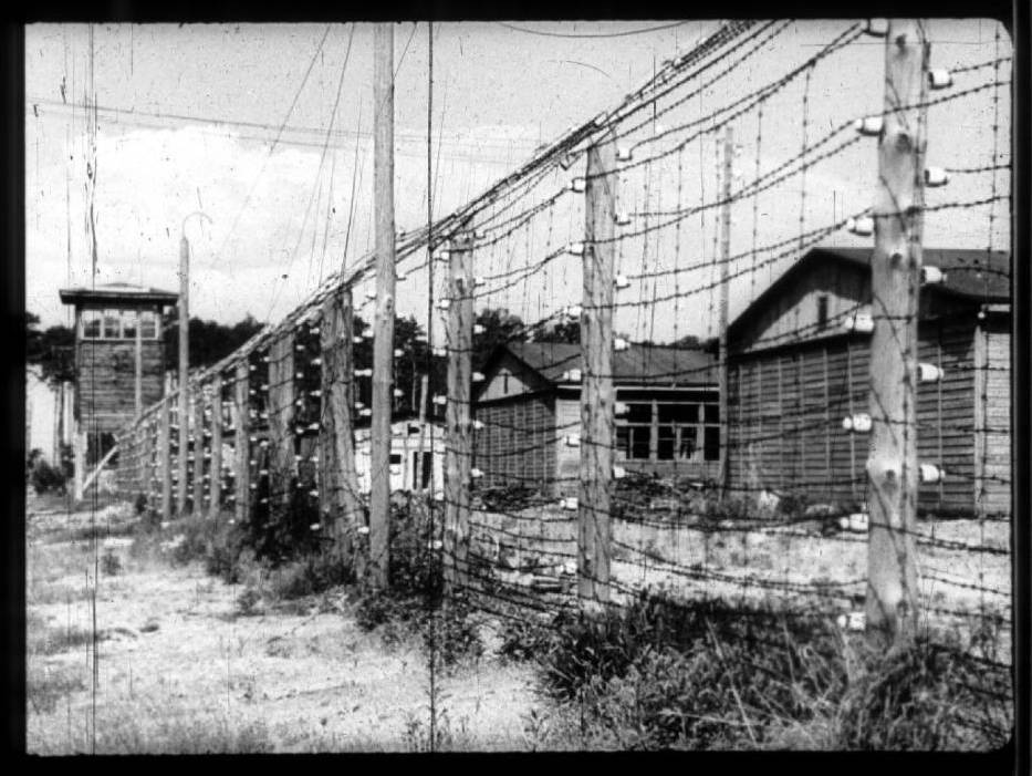 Czarno-białe zdjęcie przedstawia stan obozu w 1945 roku. Na pierwszym planie drut kolczasty i wieżyczka wartownicza. Za drutem widać zniszczone baraki. 