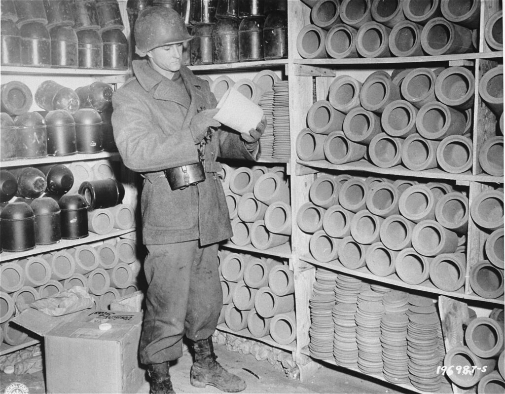 Zdjęcie czarno białe przedstawiające żołnierza amerykańskiego z czasów II wojny światowej. W otoczeniu regałów z urnami. Z lewej strony widoczne krążki szamotowe. 