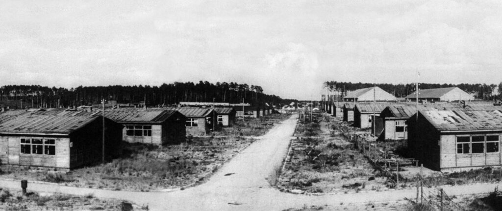 Zdjęcie czarno-białe przedstawiające zniszczone baraki obozowe. Po środku szutrowa droga. 