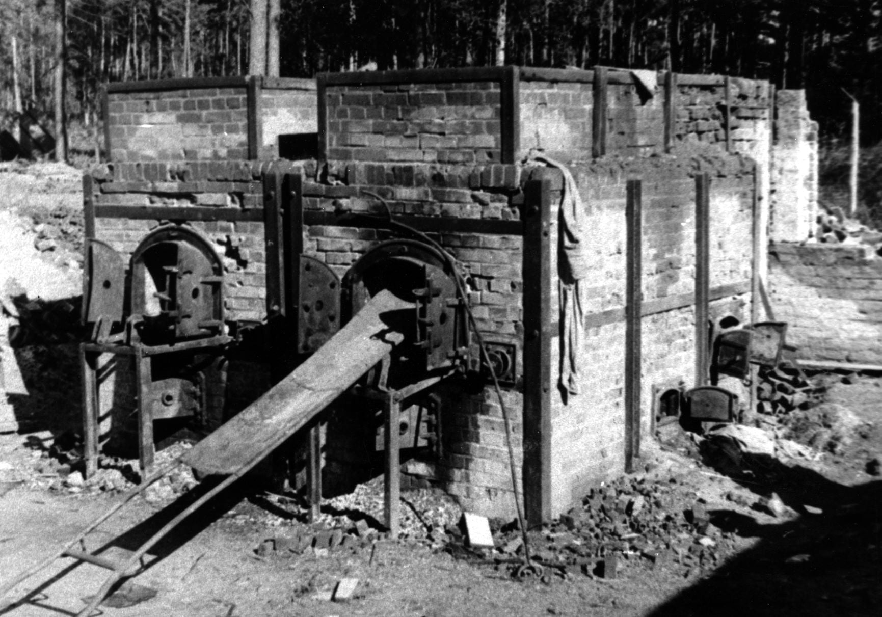 Czarno-białe zdjęcie przedstawiające ceglane piece krematoryjne. Widoczne metalowe drzwiczki oraz łóżko służące do wkładania zwłok więźniów.