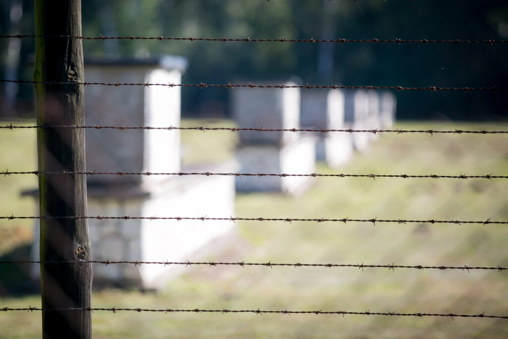 Zdjęcie przedstawia zbliżenie na ogrodzenie z drutu kolczastego z rozmytymi betonowymi pomnikami w tle. Konstrukcje są ustawione w rzędzie, a scena jest obszarem zewnętrznym z trawiastym polem i drzewami w oddali.