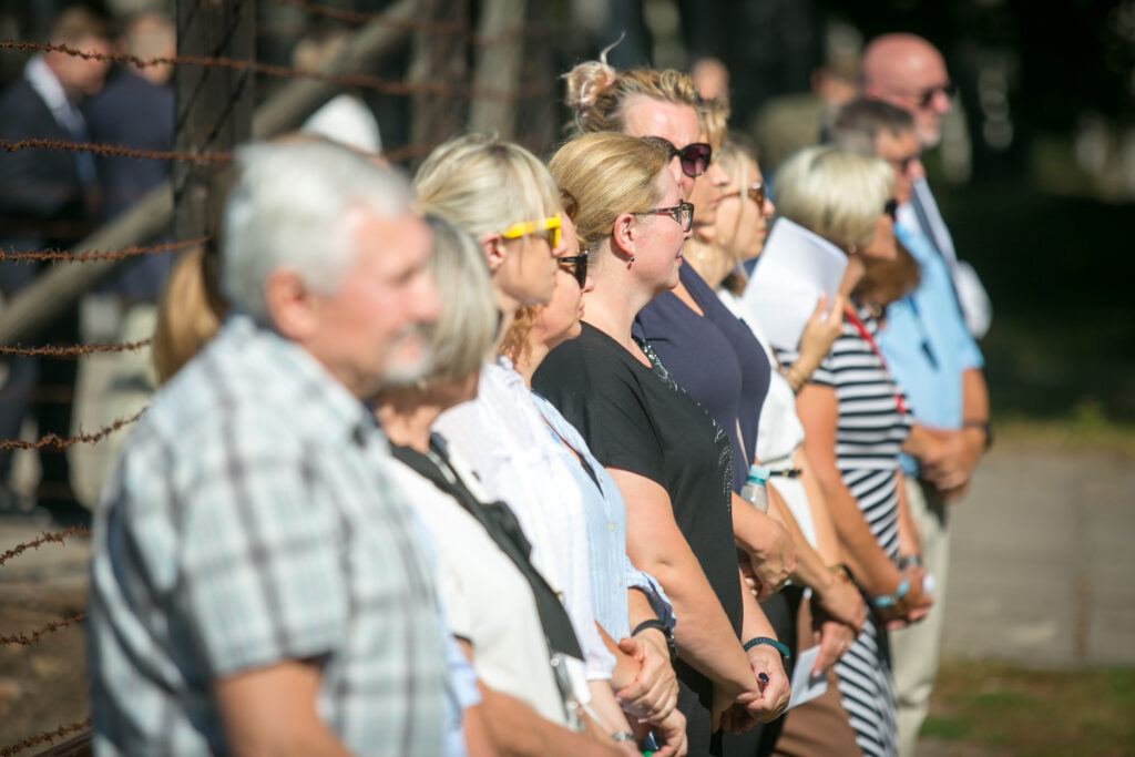 Zdjęcie przedstawia grupę ludzi stojących w rzędzie na zewnątrz, uważnie obserwując coś poza kadrem po prawej stronie. Stoją przy drucie kolczastym, a światło słoneczne rzuca cienie na ziemię. Tło jest rozmyte, widoczne są drzewa i zieleń.