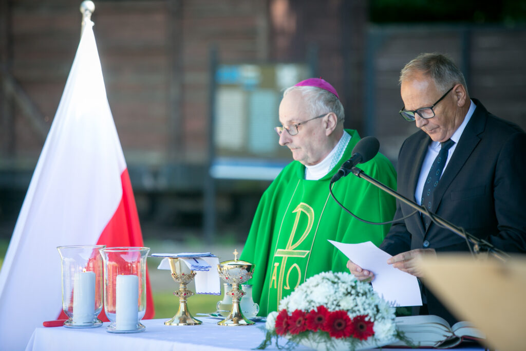 Zdjęcie przedstawia ceremonię religijną, która jest prowadzona na zewnątrz. Jeden mężczyzna, ubrany w zieloną szatę duchowną z jarmułką, stoi za ołtarzem z zapalonymi świecami, kielichem i kwiatami. Inny mężczyzna w okularach i garniturze czyta dokument. W tle widać flagę.