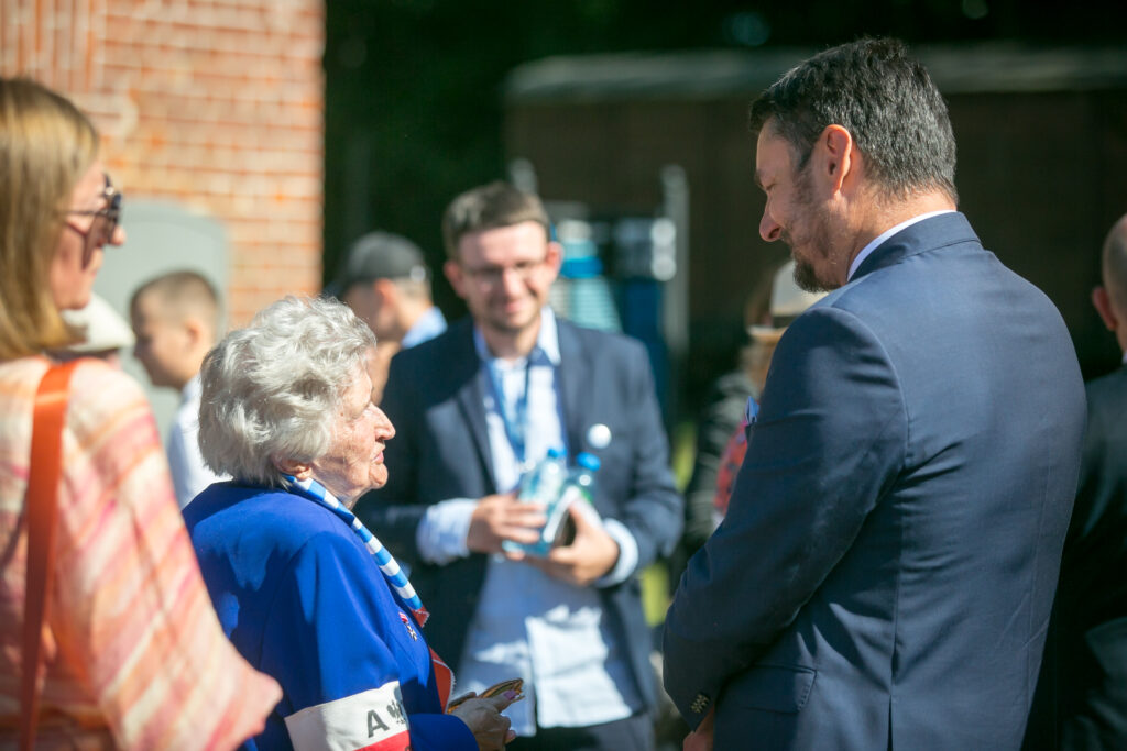 Zdjęcie przedstawia grupę ludzi, którzy stoją na zewnątrz, rozmawiając. Starsza kobieta ubrana w niebieską marynarkę i białą opaskę na ramieniu z numerem na niej, stoi w środku, rozmawiając z mężczyzną w garniturze. Inne osoby w tle, w tym mężczyzna trzymający przedmiot w rękach, słuchają uważnie.