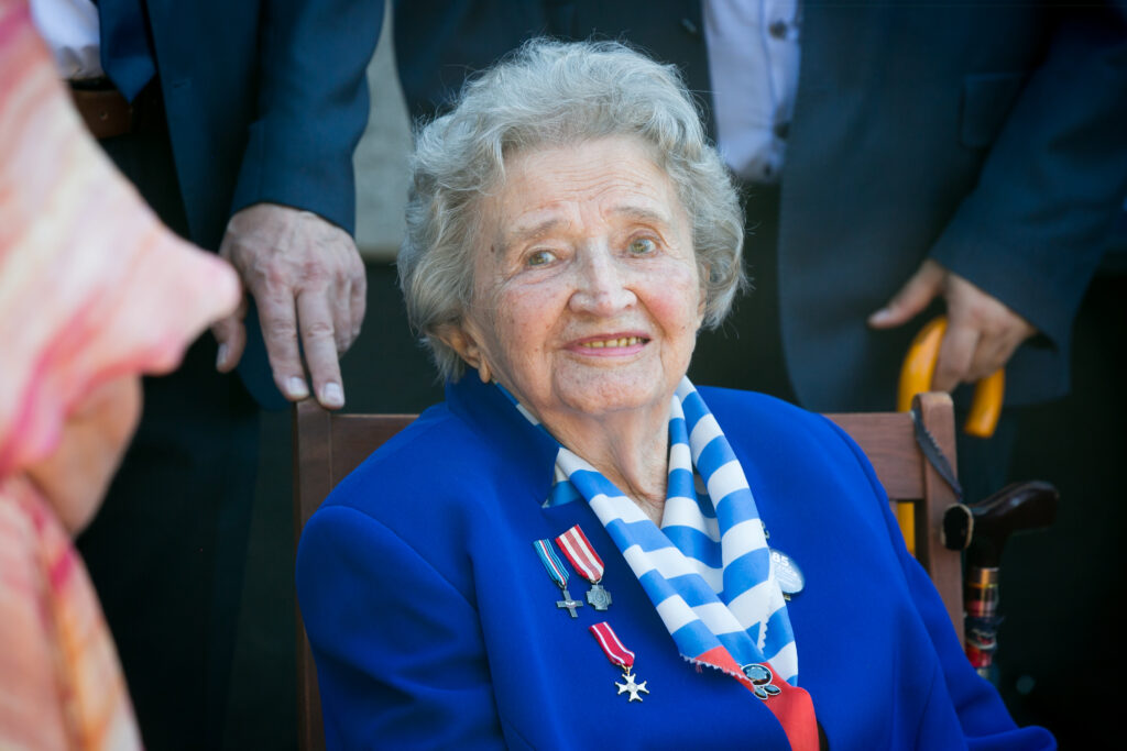 Zdjęcie przedstawia Marię Kowalską - starszą kobietę, która siedzi na krześle, ubrana w niebieską marynarkę z medalami i chustą w paski. Ma krótkie siwe włosy i się uśmiecha. Na oparciu krzesła widać dłoń osoby, a obok niej widać laskę. W tle stoją ludzie.