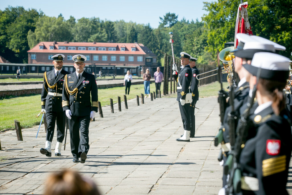 Zdjęcie przedstawia formalną ceremonię, podczas której oficer marynarki w mundurach idzie utwardzoną ścieżką. Dwóch oficerów idzie obok siebie, podczas gdy inni stoją w szyku. W słoneczny dzień w tle widać zabytkowy budynek komendantury i bujną zieleń.