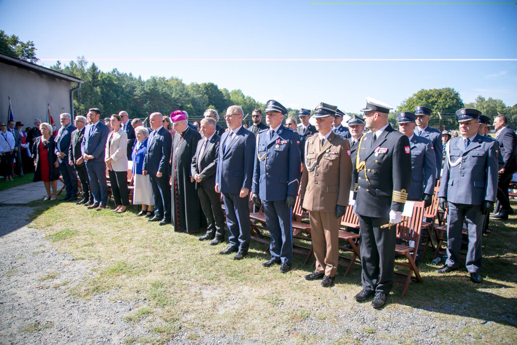 Zdjęcie przedstawia grupę ludzi, w tym personel wojskowy w różnych mundurach i cywili w formalnych strojach, która stoi na zewnątrz podczas uroczystości, które jest wydarzeniem upamiętniającym. Niektórzy mają za sobą składane, drewniane krzesłach na trawiastym terenie pod czystym, błękitnym niebem.