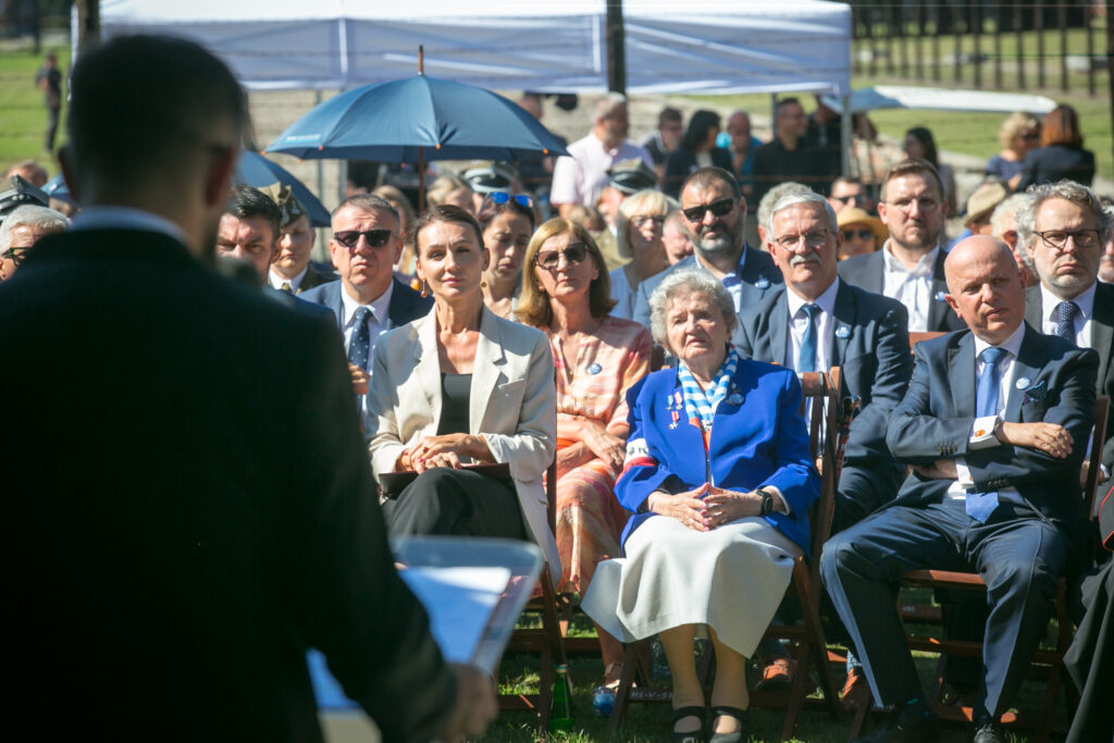 Zdjęcie przedstawia grupę osób siedzących na zewnątrz podczas wydarzenia, uważnie zwróconych w stronę mówcy, który jest częściowo widoczny na pierwszym planie. Niektórzy są ubrani w formalne stroje, jedna osoba trzyma parasol, a kilka osób wydaje się uważnie słuchać.