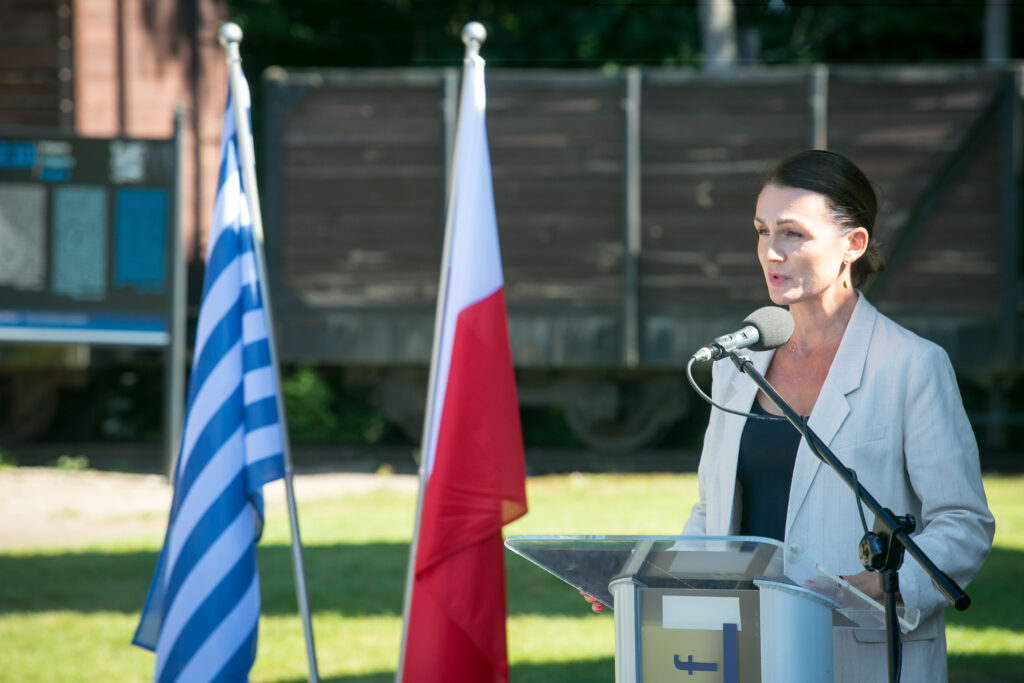 Zdjęcie przedstawia kobietę, która stoi na podium, mówiąc do mikrofonu. Za nią są dwie flagi, jedna niebiesko-biała, a druga czerwono-biała. W tle widać drewniany wagon kolejowy i drzewa. Kobieta ma na sobie szarą marynarkę i ciemne włosy związane do tyłu.