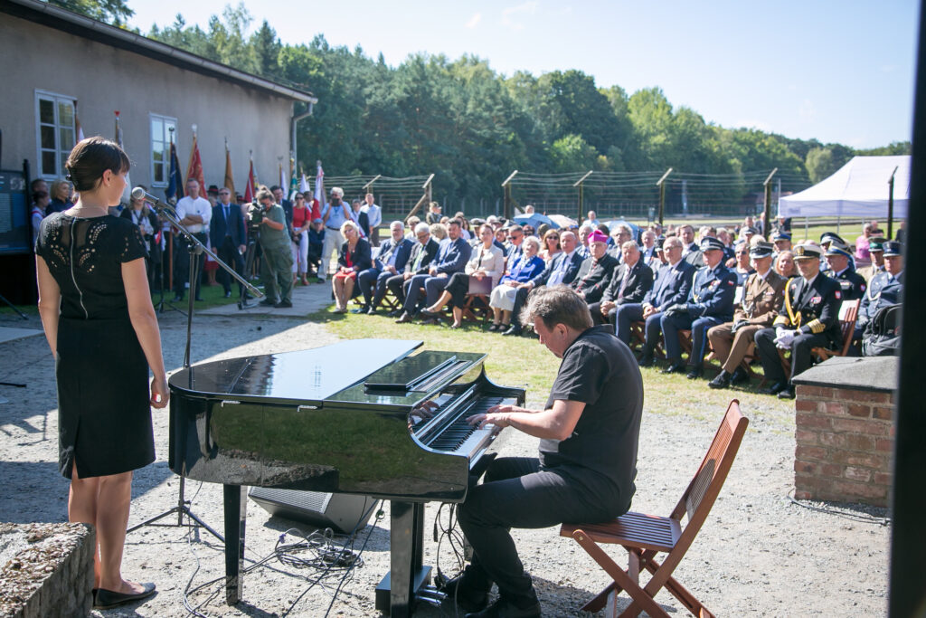 Zdjęcie przedstawia kobietę, która stoi przed mikrofonem, a mężczyzna obok niej gra na fortepianie. Stają twarzą do publiczności siedzącej w rzędach na zewnątrz, w tym kilku osób w mundurach wojskowych. Wydarzenie odbywa się w pobliżu budynku z flagami i otoczonego drzewami.