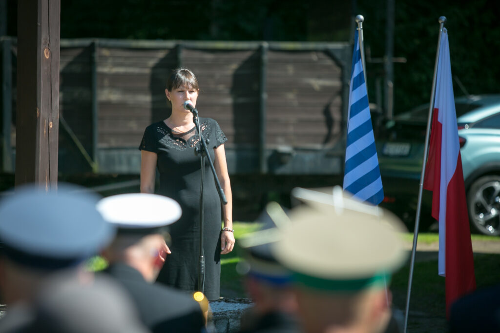 Zdjęcie przedstawia kobietę w czarnej sukience, która stoi przy mikrofonie, śpiewa na zewnątrz. Na pierwszym planie widać kilka osób w mundurach wojskowych i czapkach, zwróconych w jej stronę. Za nią widać dwie flagi, jedną z niebiesko-białymi paskami, a drugą czerwono-białą.