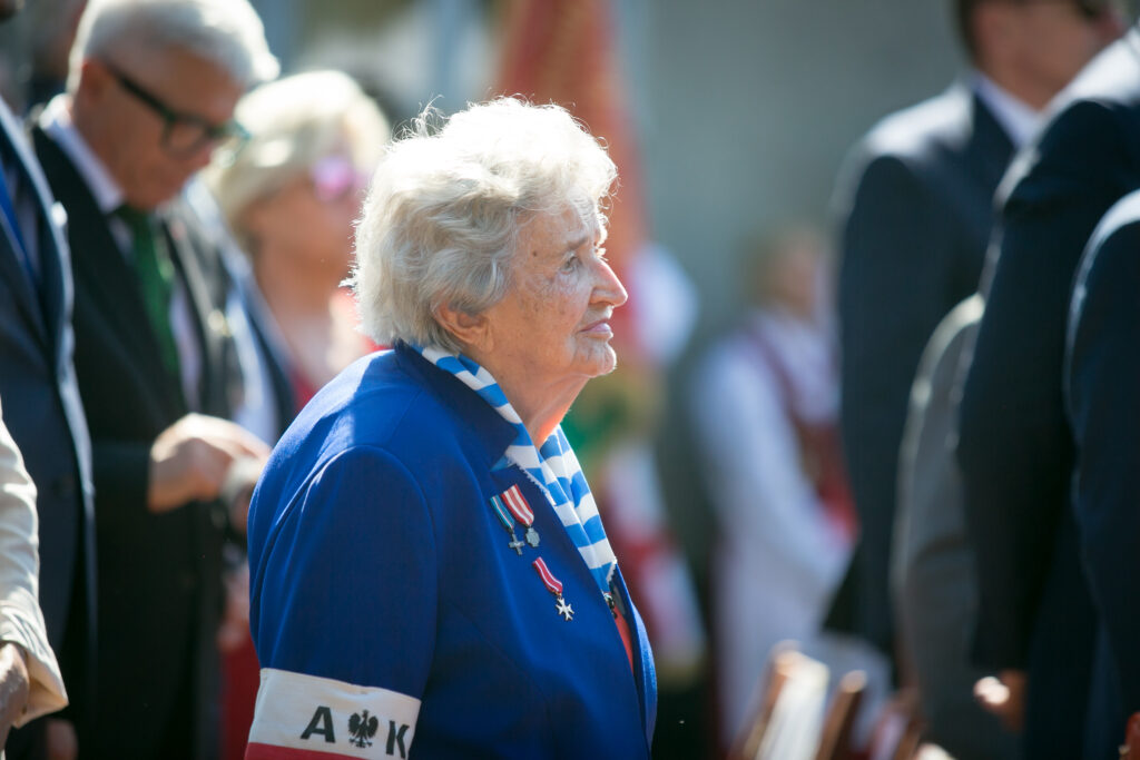 Zdjęcie przedstawia Marię Kowalską - Starszą kobietę z białymi włosami, ubraną w niebieską marynarkę ozdobioną medalami i biało-niebieską chustą, stoi ona wśród grupy ludzi. Jest na uroczystości, z niewyraźnymi postaciami w tle.