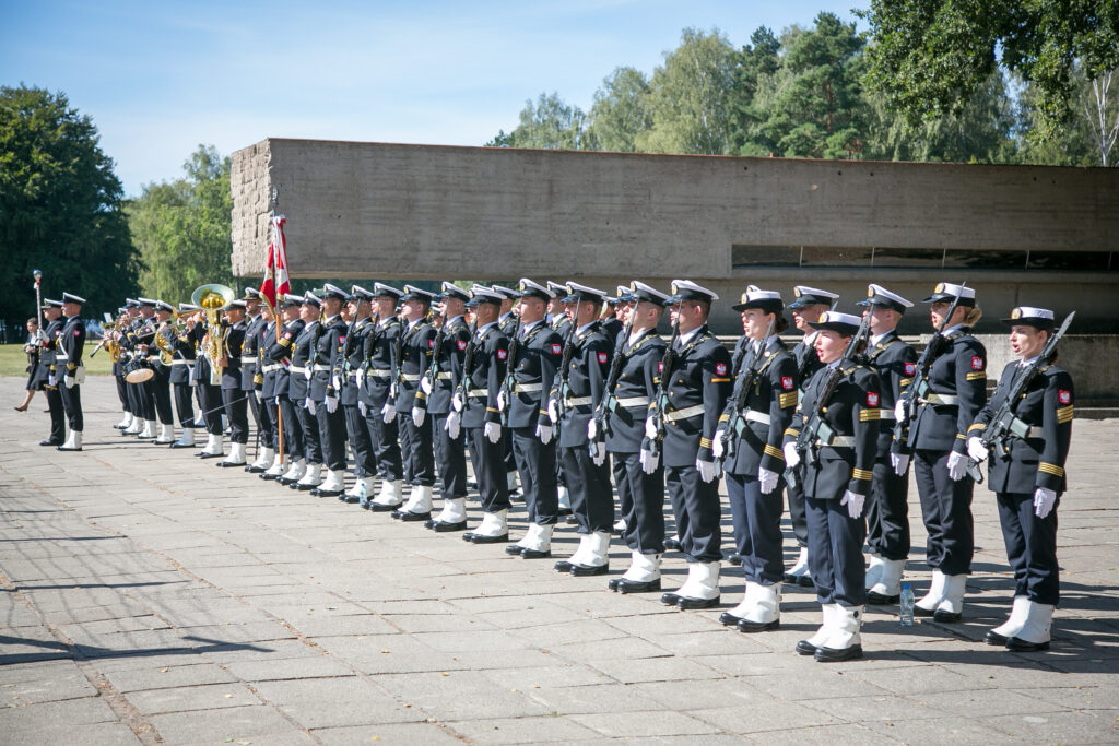 Zdjęcie przedstawia umundurowaną orkiestrę marszową, która stoi w rzędach na zewnątrz podczas ceremonii. Członkowie noszą mundury marynarki wojennej, białe rękawiczki i czapki z białymi opaskami, trzymając różne instrumenty muzyczne. W tle widać dużą betonową konstrukcję i drzewa.