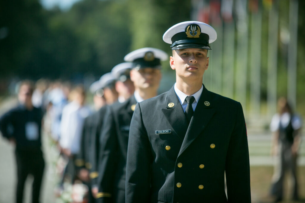 Zdjęcie przedstawia żołnierzy w formalnych mundurach marynarki wojennej, w tym czapkach, którzy maszerują w szeregu na zewnątrz. Uwaga skupiona jest na mężczyźnie z przodu, z innymi i flagami w rozmytym tle. 