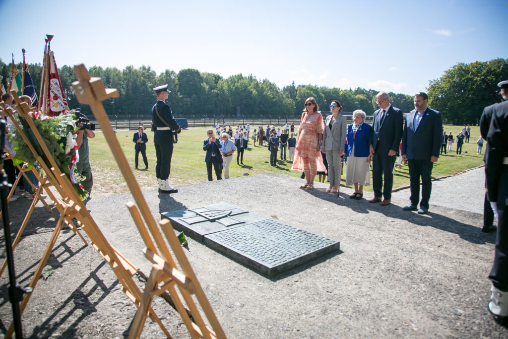 Zdjęcie przedstawia ludzi stojących wokół tablicy pamiątkowej na zewnątrz w słoneczny dzień. Różne flagi są wystawione na drewnianych stojakach w pobliżu. W tle widać drzewa i trawiaste pole, a w oddali gromadzą się kolejne osoby.