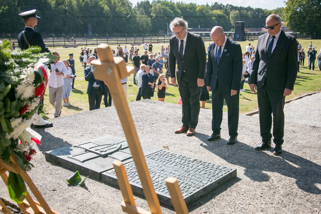 Zdjęcie przedstawia grupę ludzi stojących uroczyście wokół tablicy pamiątkowej na platformie na świeżym powietrzu. Trzech mężczyzn w formalnych strojach, w tym garniturach i krawatach, pochyla głowy z szacunkiem. Na pierwszym planie częściowo widoczny jest wieniec kwiatów. W tle zgromadził się tłum.