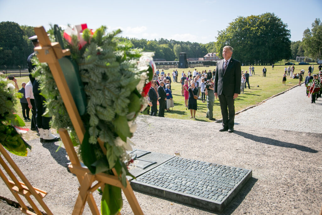 Zdjęcie przedstawia ludzi, którzy gromadzą się na zewnątrz. Na pierwszym planie widać kilka stojących wieńców, a mężczyzna w garniturze patrzy w ich stronę. W tle widać drzewa i budynek wraz z wieżą wartowniczą. Jest słoneczna pogoda podczas uroczystości. 