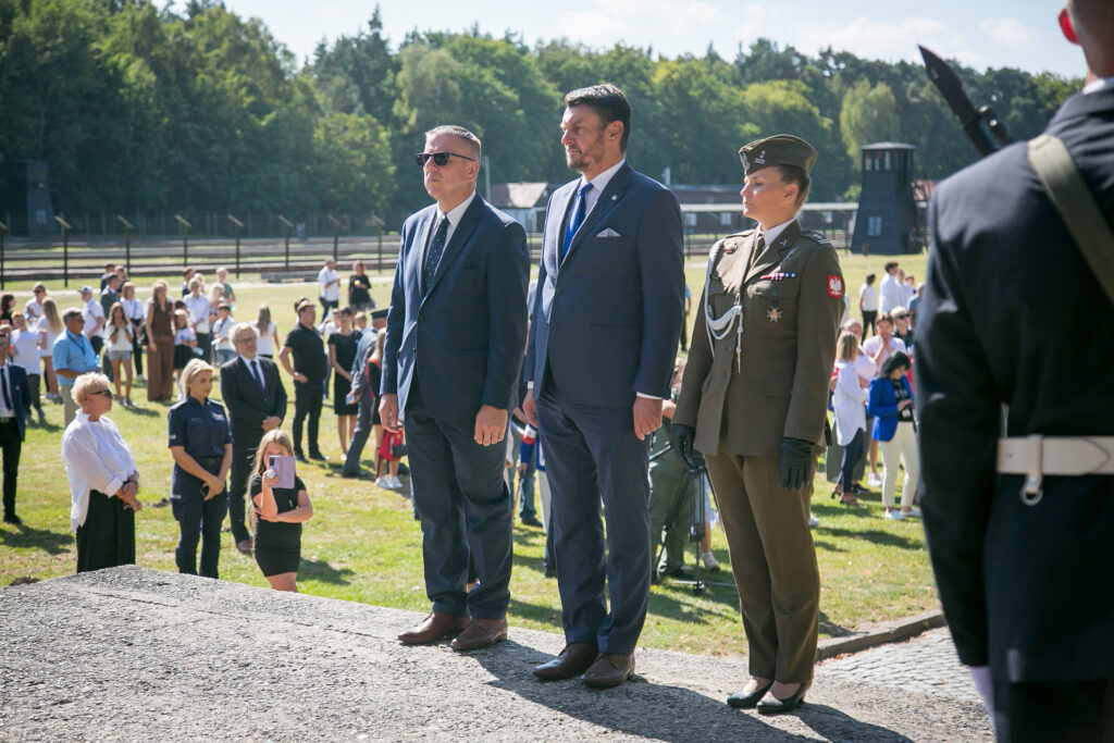 Zdjęcie przedstawia na pierwszym planie - trzy osoby, dwóch mężczyzn w garniturach i kobietę w mundurze wojskowym, stoją na platformie na zewnątrz, obok zgromadzonego tłumu. Wydarzenie jest formalne, z wieloma uczestnikami widocznymi w tle i bujnym lasem w oddali pod czystym niebem.