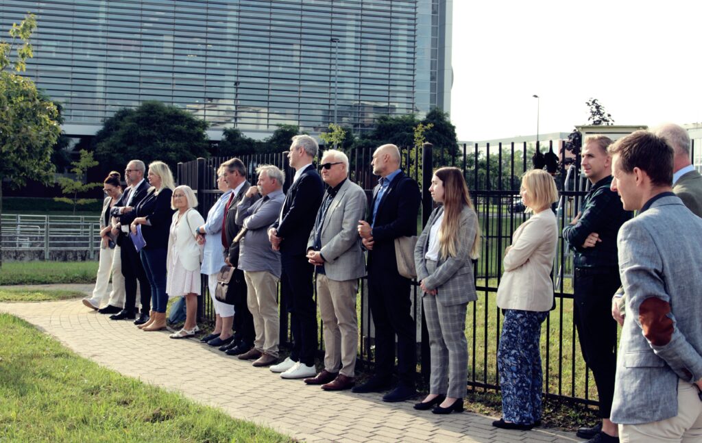 Oczywiście! Oto alternatywny opis Twojego zdjęcia:

Grupa osób stoi w linii na trawiastym terenie, z twarzami zasłoniętymi brązowymi prostokątami dla zachowania prywatności. W tle znajduje się nowoczesny budynek z refleksyjnymi szklanymi oknami. Sceneria sugeruje, że jest to miejskie otoczenie, prawdopodobnie wskazujące na formalne spotkanie lub wydarzenie na zewnątrz budynku biurowego.
