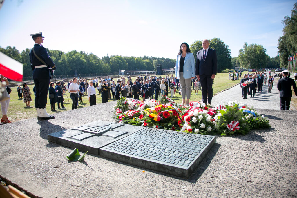 Zdjęcie przedstawia ludzi, którzy stoją uroczyście wokół pomnika z wieńcami i kwiatami, podczas gdy osoby w formalnych strojach, w tym żołnierz ubrany w mundur, są obecne. Drzewa i tłum w tle podkreślają wydarzenie, które jest ceremonią. 