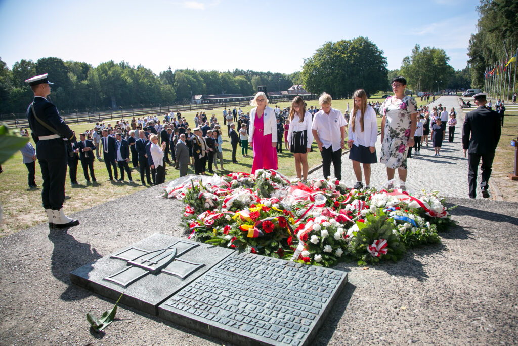 Zdjęcie przedstawia grupę ludzi, którzy zebrali się w miejscu pamięci, składając wieńce i kwiaty na kamiennym pomniku. Grupa stoi na pierwszym planie, oddając hołd, podczas gdy większy tłum jest widoczny w tle. Obok nich stoi żołnierz patrzący w stronę ludu. Drzewa i maszt flagowy z flagami są widoczne pod jasnym, czystym niebem.