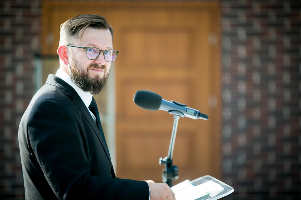 Zdjęcie przedstawia mężczyznę w okularach, garniturze i z brodą, który przemawia do mikrofonu na statywie. Stoi w budynku przed drewnianymi drzwiami i ceglaną ścianą, trzymając przed sobą kartki na mównicy. Na jego twarzy maluje się lekki uśmiech.