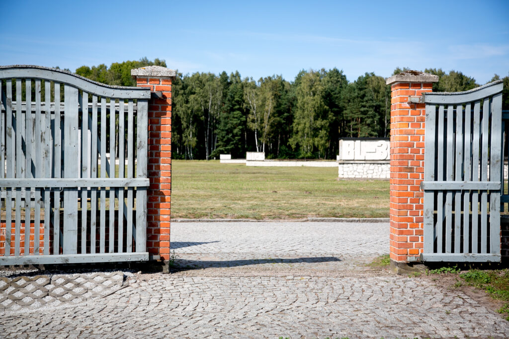 Zdjęcie przedstawia otwartą, szarą drewnianą bramę, która jest osadzona w murowanych filarach z czerwonej cegły. Brama prowadzi do trawiastej przestrzeni z białymi kamiennymi obiektami, które są pomnikiem po dawnych barakach w KL Stutthof i drzewami w tle.