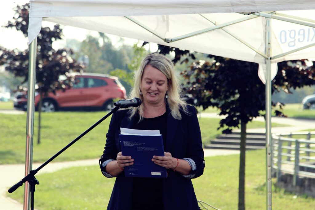 Na zdjęciu widzimy osobę stojącą pod białym zadaszeniem, trzymającą w rękach coś, co wygląda na broszurę lub dokumenty z tekstem. W tle widać zieleń oraz czerwony samochód