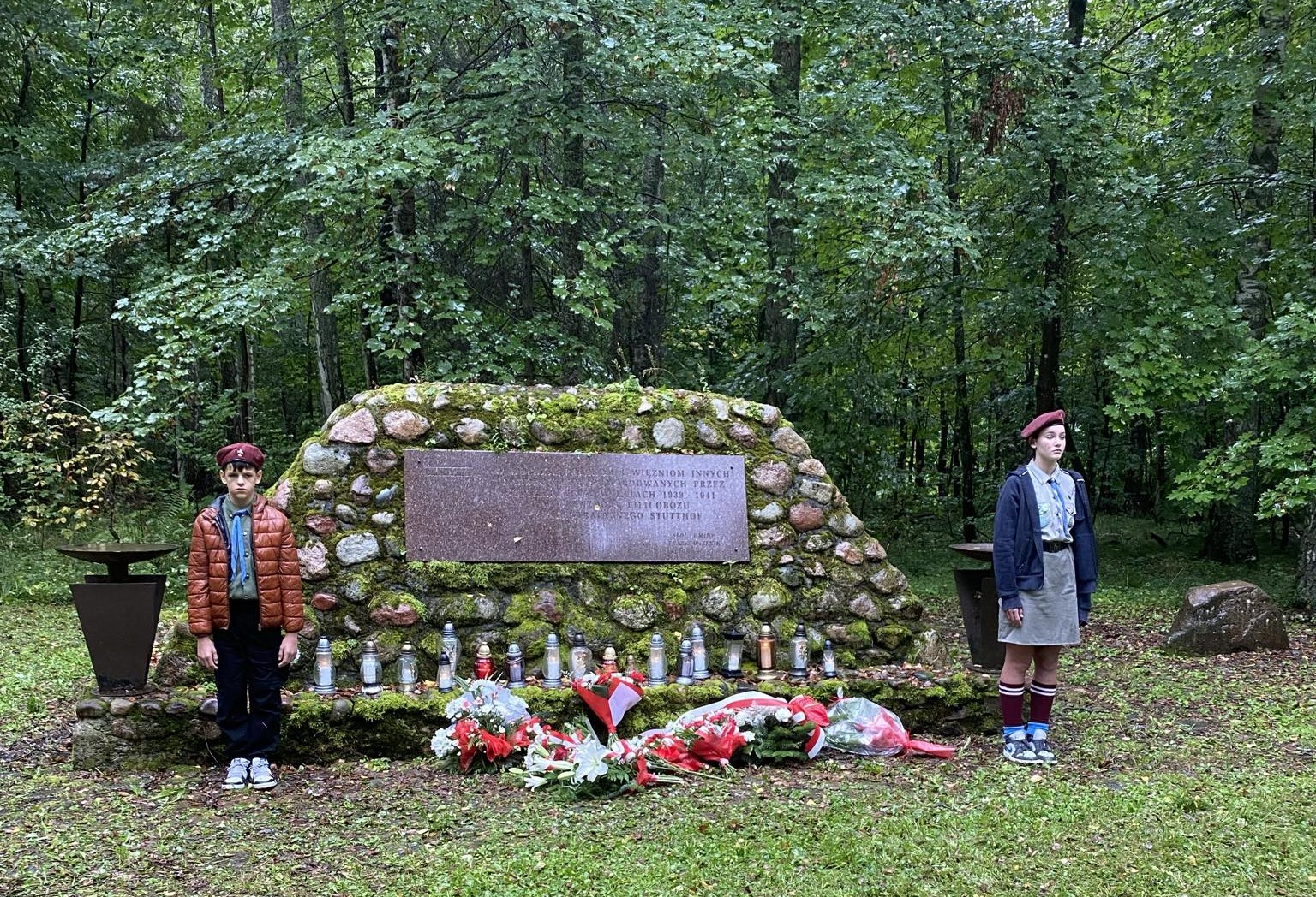 Dwoje dzieci w harcerskich mundurach stoi po obu stronach pomnika. Pomnik wykonany jest z kamieni i zawiera tablicę pamiątkową. Przed nim znajdują się znicze oraz liczne wieńce kwiatowe. W tle widoczne są drzewa w lesie, a trawa pokrywa teren wokół pomnika.