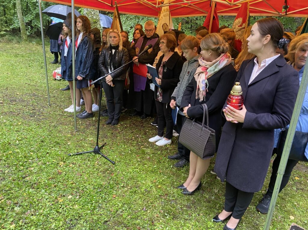 Kilkanaście osób, w tym młodzież i dorośli, stoi pod namiotem podczas uroczystości. Niektóre osoby trzymają flagi lub znicze, a mikrofon ustawiony jest przed grupą. Wszyscy są ubrani ciepło ze względu na pogodę, co wskazuje na chłodny i deszczowy dzień.