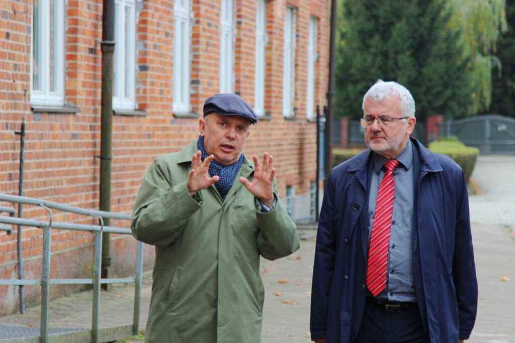  Dwóch mężczyzn spaceruje obok ceglanego budynku. Jeden z nich, ubrany w zieloną kurtkę i czapkę, gestykuluje, jakby coś wyjaśniał. Drugi mężczyzna w ciemnym płaszczu i czerwonym krawacie słucha z uwagą. Tło to ceglane ściany budynku i chodnik.