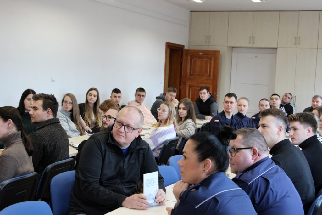 Grupa młodych osób, w tym studentów Akademii Marynarki Wojennej, siedzi w sali konferencyjnej i słucha prelekcji. Widać różnorodne reakcje – niektórzy uczestnicy spoglądają na prowadzących, inni w stronę materiałów na stole.