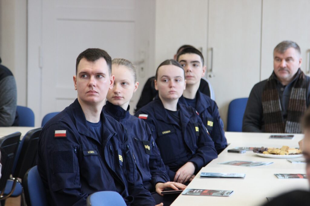 Czterech studentów w mundurach Akademii Marynarki Wojennej siedzi przy stole konferencyjnym. Ich twarze wyrażają skupienie. W tle widoczne są materiały edukacyjne, a na stole stoi talerz z przekąskami.