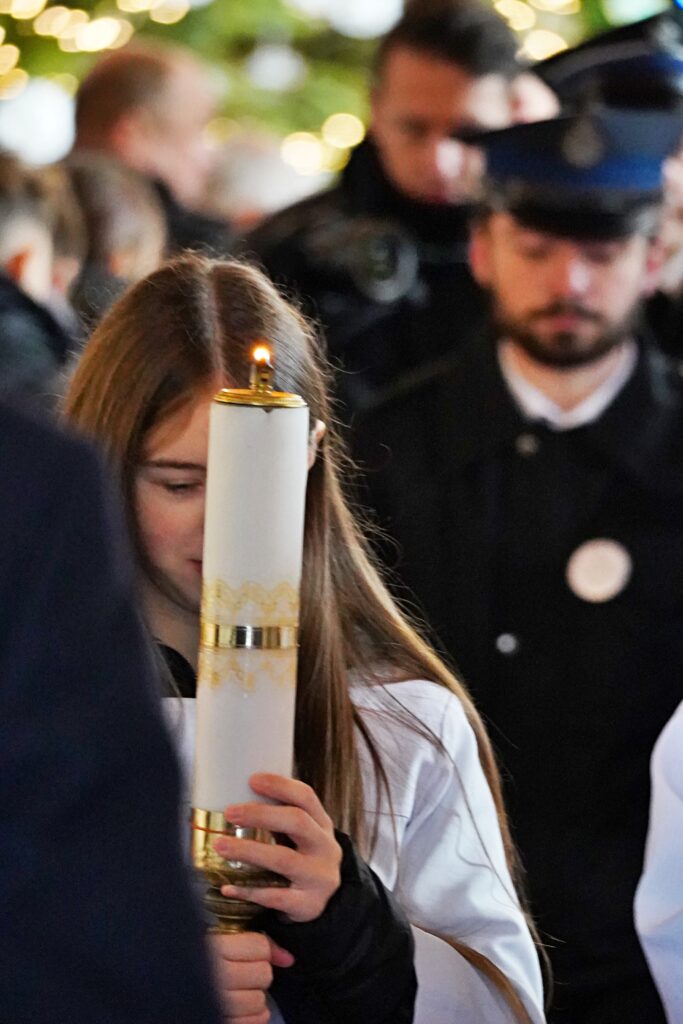 "Młoda dziewczyna w białej albie trzyma zapaloną, dużą świecę podczas uroczystości. W tle widać rozmyte sylwetki innych uczestników, w tym mężczyznę w mundurze strażackim."