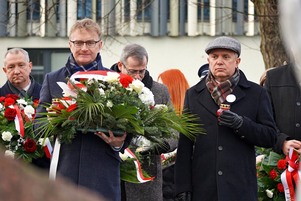 "Grupa mężczyzn podczas ceremonii składania wieńców. Jeden z nich trzyma wieniec z biało-czerwonymi kwiatami, a inny znicz. W tle widać innych uczestników i fragment budynku."