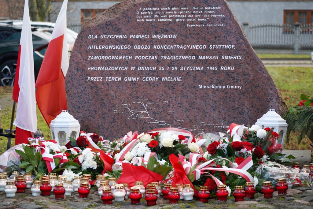 "Pomnik w kształcie płyty z czerwonego granitu z wyrytym napisem upamiętniającym ofiary Marszu Śmierci w 1945 roku. Przed pomnikiem znajdują się biało-czerwone kwiaty, wstęgi oraz zapalone znicze. Obok widoczne są flagi Polski."