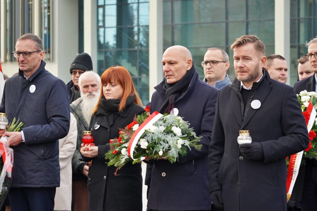 "Uczestnicy ceremonii z wieńcami i zniczami stoją w skupieniu. Na pierwszym planie kobieta z czerwonymi włosami trzyma wieniec z biało-czerwonymi kwiatami, za nią stoi starszy mężczyzna z brodą."