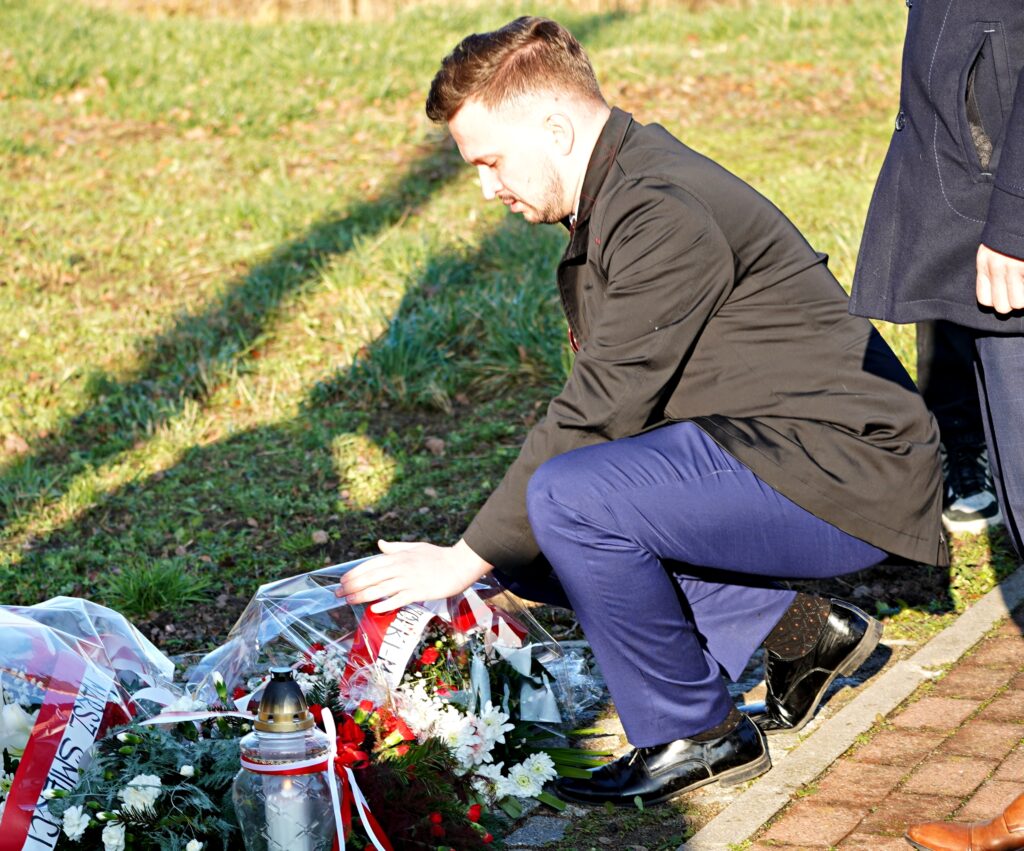 Mężczyzna w eleganckim stroju kuca, składając wieniec z biało-czerwonymi kwiatami i wstęgą podczas uroczystości. Obok na ziemi widoczne są inne wieńce i zapalone znicze. 