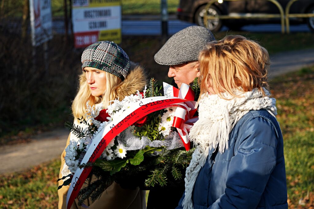 Trzy osoby, dwie kobiety i mężczyzna, stoją w skupieniu podczas ceremonii. Jedna z kobiet trzyma wieniec z biało-czerwonymi kwiatami z wstęgą. W tle widoczne są tablice reklamowe.