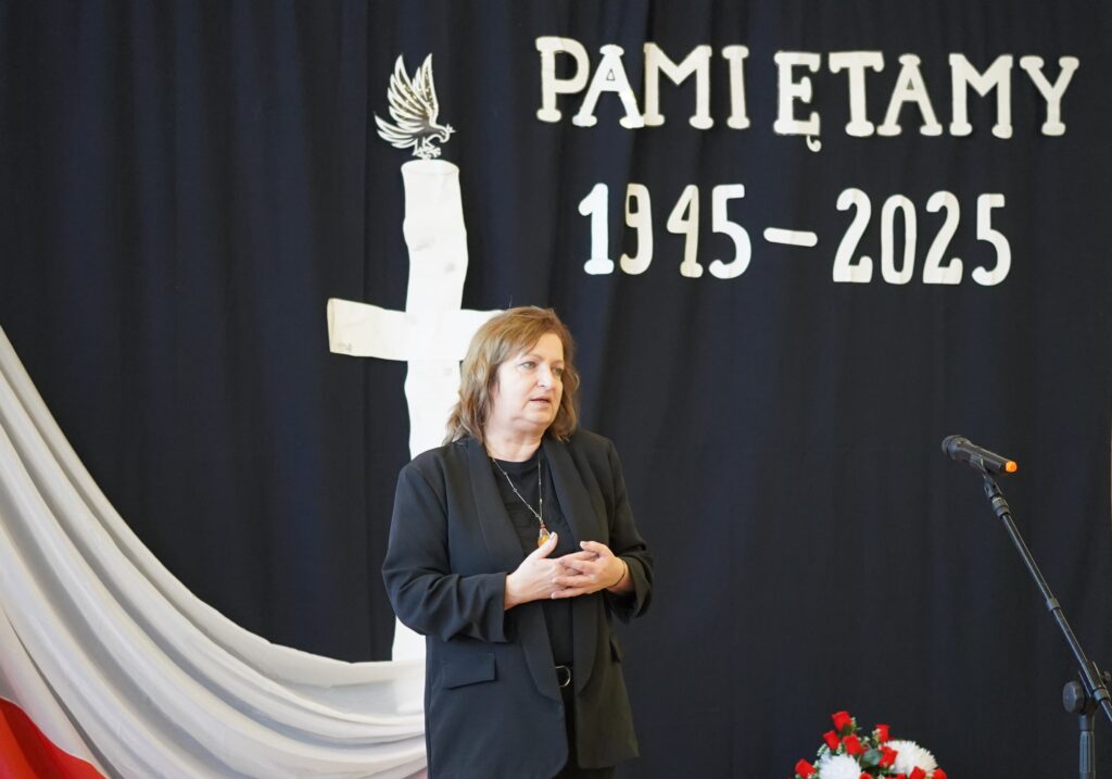 Kobieta w ciemnym stroju stoi na tle dekoracji z biało-czerwonych materiałów oraz napisu 'Pamiętamy 1945-2025'. W tle widać biały krzyż i symbol orła.