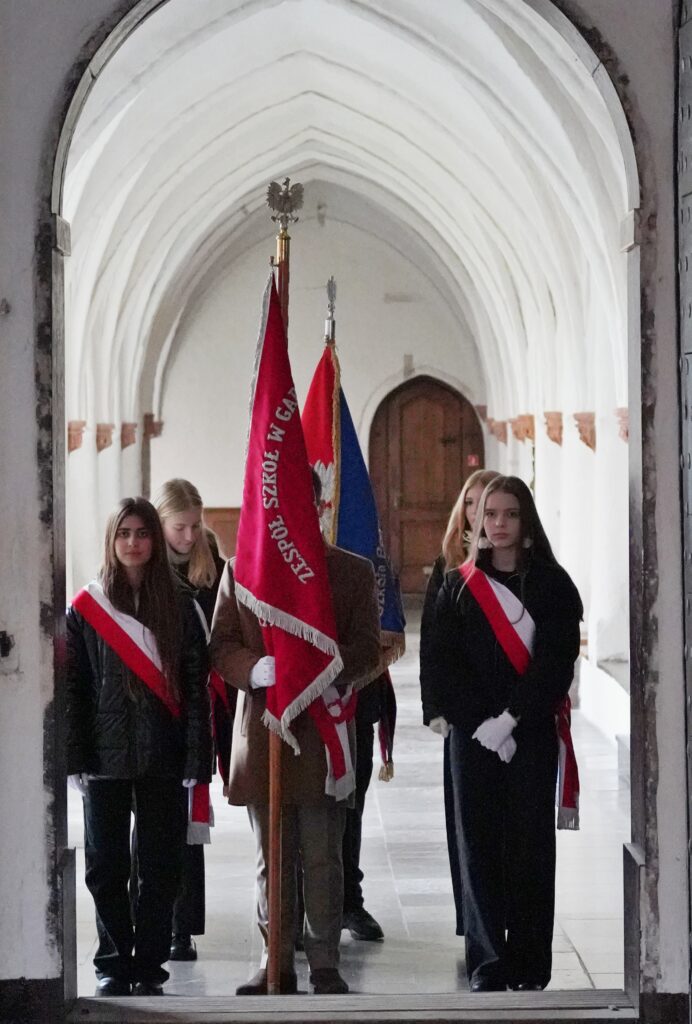 "Grupa uczniów w ceremonialnych strojach trzymających sztandary. Znajdują się w gotyckim korytarzu z widocznym łukowym sklepieniem i drzwiami w tle."